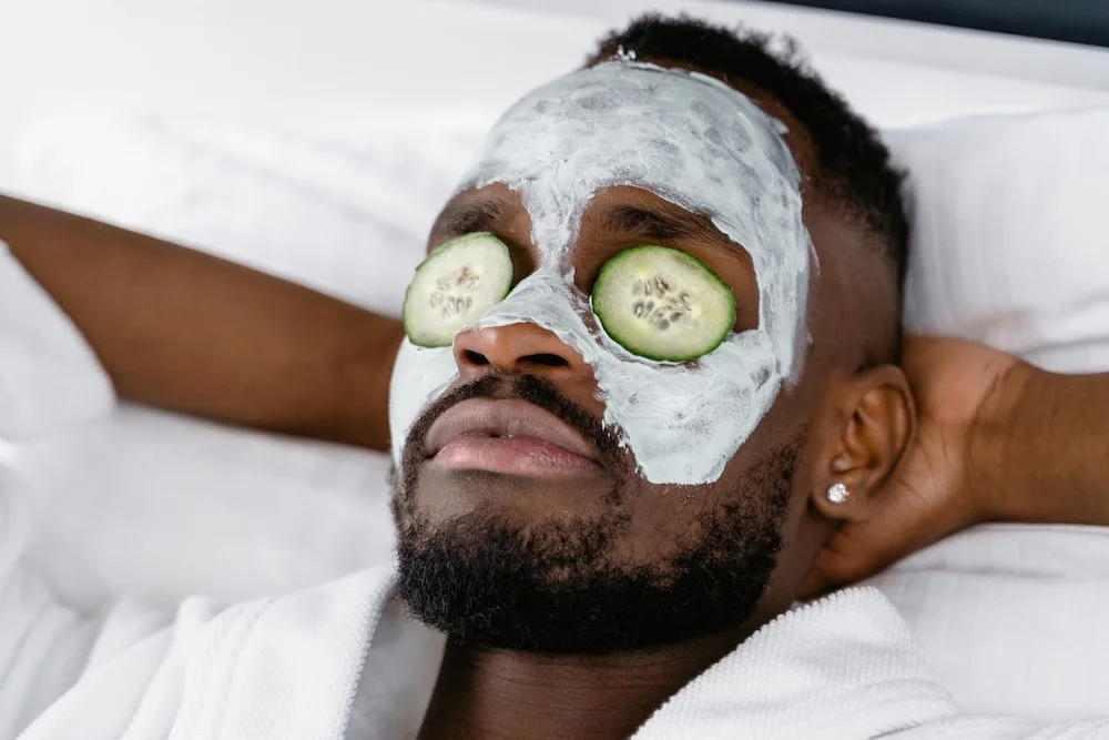 Pessoa deitada fazendo tratamento de pele com creme e rodelas de pepino sobre os olhos. A pessoa tem barba escura e cabelo curto e apresenta expressão relaxada enquanto cruza os dedos das mãos atrás da cabeça.