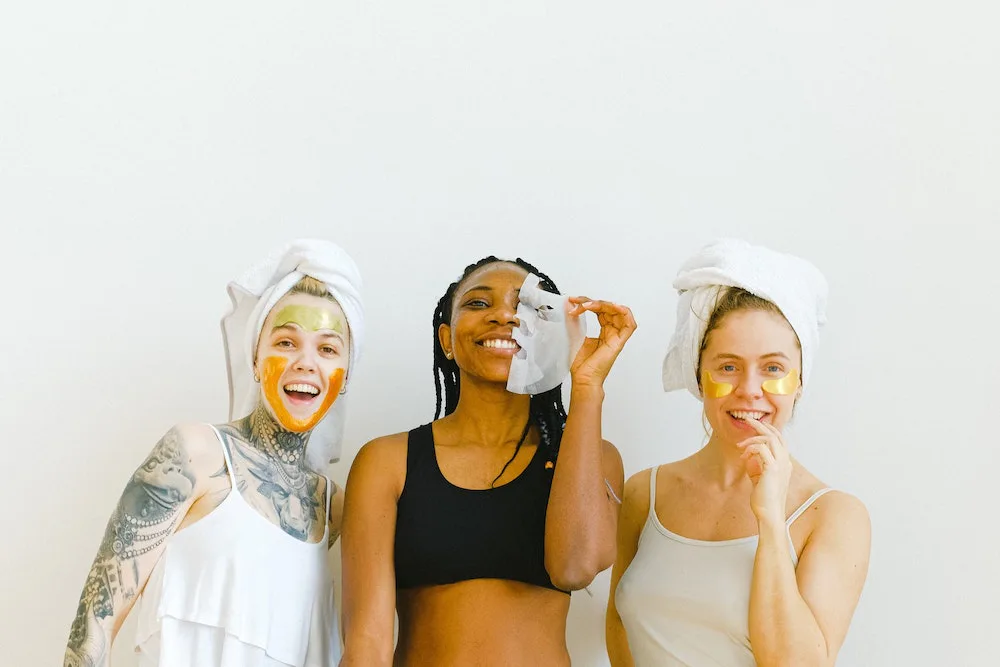 Tres pessoas lado a lado usando produtos de skincare enquanto sorriem. A pessoa do meio tira uma mascara facil do rosto. As pessoas nas laterais estão usando produtos no rosto e toalha enrolada no cabelo.
